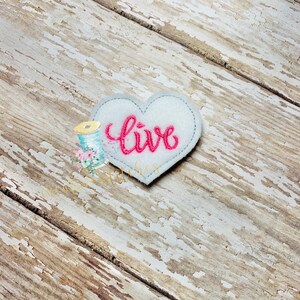 Pode incluir: Um coração de feltro branco com a palavra "live" bordada em rosa.