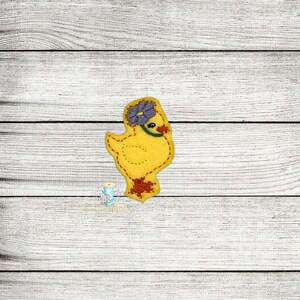 Peut inclure: Un poussin jaune en feutre avec une fleur violette sur la tête. Le poussin est debout sur un fond en bois blanc.