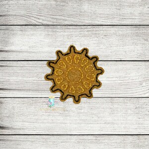 May include: A tan-colored, sun-shaped applique with a black outline. The applique has a textured appearance and a series of oval-shaped cutouts around the center. The background is a light-colored, wooden surface.