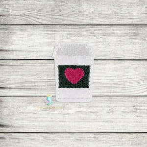 May include: White coffee cup with a green square and a pink heart in the center. The cup is on a white wood background.