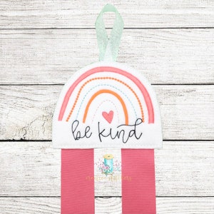 Może przedstawiać: Biały filcowy uchwyt na wstążki w kształcie tęczy z napisem "be kind" i różowym sercem. Tęcza ma pomarańczowe, brzoskwiniowe i niebieskie paski. Uchwyt ma różową wstążkę.