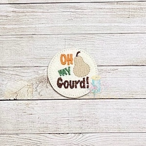 May include: A round white felt patch with brown stitching. The patch has a green and orange embroidered text that says "Oh My Gourd!" and a brown embroidered pear.