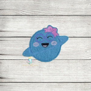 May include: A blue felt applique of a smiling planet with a pink flower on top and wearing glasses. The planet has a white ring around it.