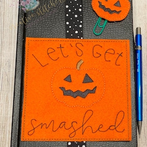 May include: Orange felt bookmark with the text "Let's get smashed" and a black felt pumpkin with a jack-o'-lantern face. The bookmark is attached to a black journal with white polka dots.