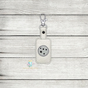May include: A white leather keychain with a gray circle design. The keychain has a silver metal ring and a silver metal clip.