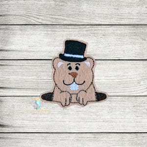 May include: Embroidered felt patch of a brown groundhog wearing a black top hat with a white band. The groundhog is sitting with its paws in front of it and has a white mouth.