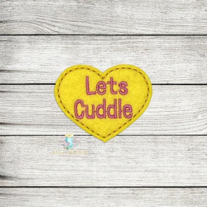 May include: A yellow felt heart with pink embroidery that reads "Lets Cuddle".