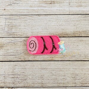 May include: Pink felt sleeping bag applique with brown stitching. The sleeping bag is rolled up and has a brown strap.