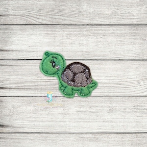 Pode incluir: Uma aplicação de feltro verde de uma tartaruga de desenho animado com uma concha marrom. A tartaruga tem um olho grande e uma boca pequena.