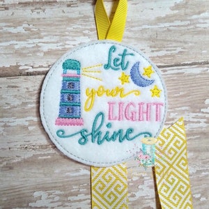 May include: A white felt circle with a blue and pink lighthouse, yellow stars, a blue crescent moon, and the text "Let your light shine" in pink and teal embroidery.
