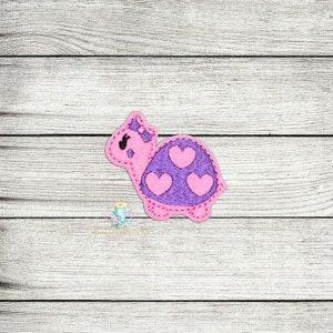 May include: An embroidered turtle with a pink and purple shell, featuring three pink hearts.