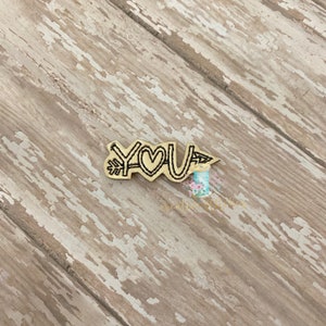 May include: A gold-colored wooden cutout with a black outline of an arrow with the word "YOU" and a heart in the middle.