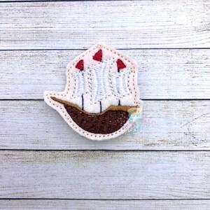 May include: A white felt applique of a ship with three red sails and a brown hull. The ship is outlined in brown thread.