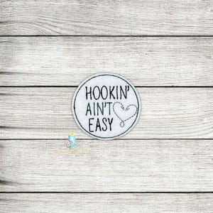 Puede incluir: Parche redondo blanco bordado con el texto "Hookin' Ain't Easy" y un pequeño corazón con un anzuelo dentro.