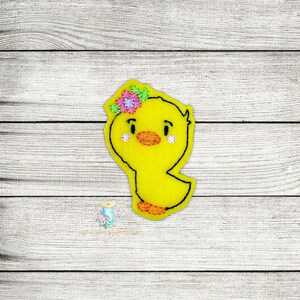 Peut inclure: Une applique en feutre jaune représentant un poussin avec une couronne de fleurs rose et verte. Le poussin a des yeux noirs, un bec jaune et des joues roses.