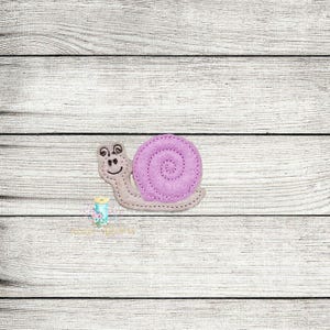 Puede incluir: Aplicación de fieltro de un caracol con concha lavanda y cara sonriente. El cuerpo del caracol es gris claro y la concha presenta un diseño en espiral. La aplicación está cosida con hilo en contraste.