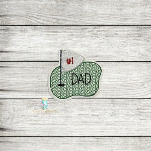 May include: Embroidered green golf green with a white flag and the text "#1 DAD" in red and white thread.