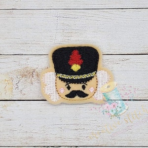 Könnte beinhalten: Gestickter Filz-Nussknacker mit schwarzem Hut, weißem Bart und braunem Schnurrbart. Der Nussknacker hat eine rote und gelbe Feder an seinem Hut.