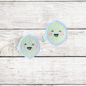 Op de afbeelding: Twee geborduurde patches in de vorm van een blad met lachende gezichten. De patches zijn lichtgroen met een lichtblauwe rand en roze wangen. De achtergrond is een wit houten oppervlak.