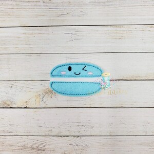 May include: A light blue felt applique of a smiling macaron with a white filling. The macaron has a winking eye and a small pink blush on each cheek.