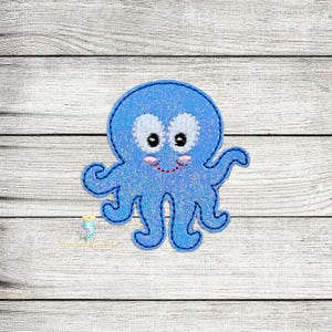 Peut inclure: Une pieuvre en feutre bleu avec un visage souriant et des yeux blancs. La pieuvre a huit tentacules et est brodée avec une finition scintillante.