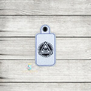 May include: White key chain with a black embroidered all-seeing eye inside a triangle with rays of light.