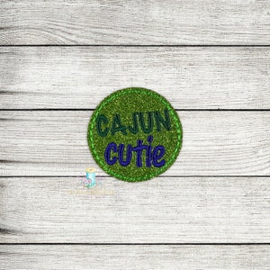 May include: Green glitter circle with the text "CAJUN cutie" in purple thread.