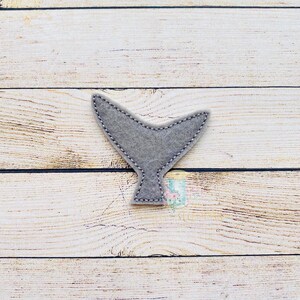 May include: A gray felt whale tail applique. The tail is a simple shape with a rounded tip and a slightly curved bottom edge.
