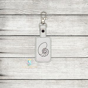 Può includere: Portachiavi in ​​pelle bianca con un portachiavi argentato e un contorno nero di una conchiglia.