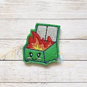 May include: A green felt patch featuring a smiling dumpster on fire. The dumpster has a white lid and is filled with red, orange, and yellow flames. The patch is embroidered with black and blue thread.