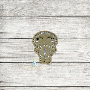 May include: A brown gingerbread person with a smiling face and white icing details. The gingerbread person is standing on a white wooden background.