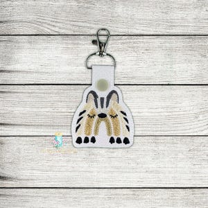 Pode incluir: Chaveiro branco com um design decorativo de duas raposas estilizadas dormindo. As raposas são em tons de bege, preto e marrom. O chaveiro tem um fecho prateado e um verso de couro branco.