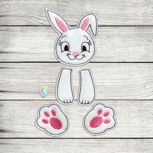 May include: A white felt bunny rabbit applique with pink ears, eyes, and nose. The bunny is separated into five pieces: head, two legs, and two feet. The feet have pink paw prints.