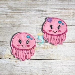 May include: Two pink felt jellyfish with embroidered faces and blue accents. The jellyfish have a smiling expression and are decorated with small blue dots.