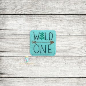 Puede incluir: Un parche de fieltro azul claro con las palabras "WILD ONE" bordadas en hilo azul oscuro. Un pequeño árbol verde está bordado entre las palabras "WILD" y una flecha marrón está bordada entre las palabras "WILD" y "ONE".