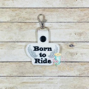 Puede incluir: Llavero de tela blanca con un clip de metal plateado. El llavero tiene un diseño bordado en negro de alas y el texto "Born to Ride".