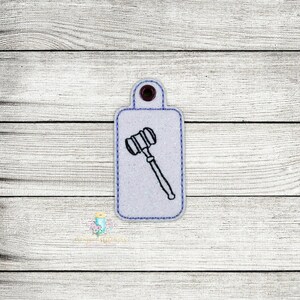 May include: A white rectangular tag with a purple border and a black gavel design. The tag has a small hole at the top with a purple grommet. The background is a light wood grain.