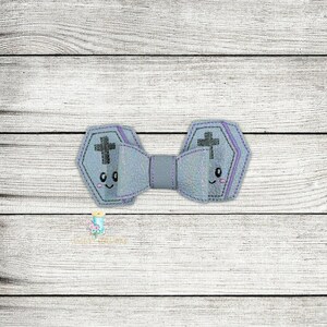 May include: A gray and purple bow tie with a cross and a cute face on each side. The bow tie is made of fabric and has a glittery finish.