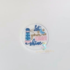 May include: A white felt circle with embroidered text that reads "Let your light shine". The design features a blue and pink lighthouse with a light beam, a blue bird, and a crescent moon.