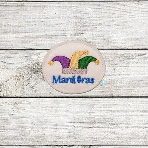 May include: A white felt patch with a colourful jester's hat and the text "Mardi Gras" in blue embroidery.