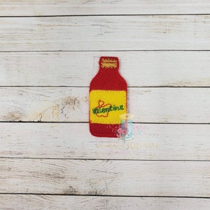 May include: A red felt bottle with a yellow label featuring the word "Valentina" in green script. The bottle has a gold-colored top and is set against a light wood grain background. The design is likely a decorative item or applique.