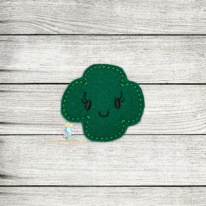 Puede incluir: Un trébol de fieltro verde con una cara sonriente y pestañas.