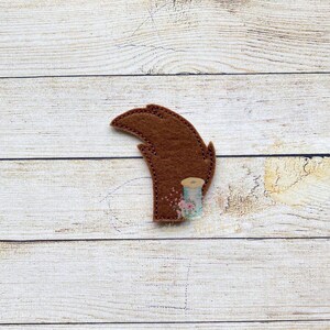 May include: A brown felt feather-shaped applique with stitched detailing. A small spool of thread with floral accents is positioned below the feather. The applique is set against a light wood grain background.