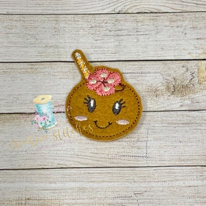 Pode incluir: Uma aplicação de feltro marrom de um rosto de desenho animado sorridente com uma flor rosa no topo e um chifre amarelo e branco.