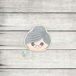 Könnte beinhalten: Gestickter Aufnäher mit dem Gesicht einer Frau mit grauem Haar, Brille und einem Dutt. Das Gesicht lächelt.