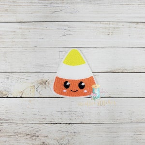 May include: A felt applique of a candy corn with a cute cartoon face. The candy corn is yellow, white, and orange.