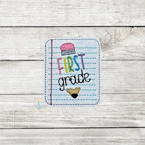 May include: A notebook-shaped patch with the words "FIRST grade" in colorful letters. A pink pencil eraser tops the design, and a small, brown pencil tip is at the bottom. The patch is on a light blue background with lines.