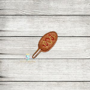 Może przedstawiać: Brązowy corndog z filcu z żółtą musztardą i czerwonym ketchupem wyszywanym na białym drewnianym tle.