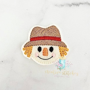 Może przedstawiać: Haftowana naszywka przedstawiająca uśmiechniętą twarz stracha na wróble, w słomkowym kapeluszu z czerwoną opaską. Strach na wróble ma czarne oczy, pomarańczowy, wyszywany nos i różowe policzki. Naszywka ma słomiane włosy i znajduje się na białym tle.