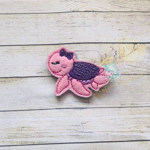 Puede incluir: Una tortuga de fieltro rosa con un caparazón morado y un lazo rosa. La tortuga tiene una cara sonriente y los ojos cerrados.